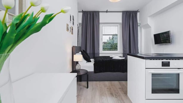 A modern, compact room with a bed, bedside table, lamp, and a small kitchen area including a stove. - Bright Studio in the Center | Close to Kleparski Park (Kraków)
