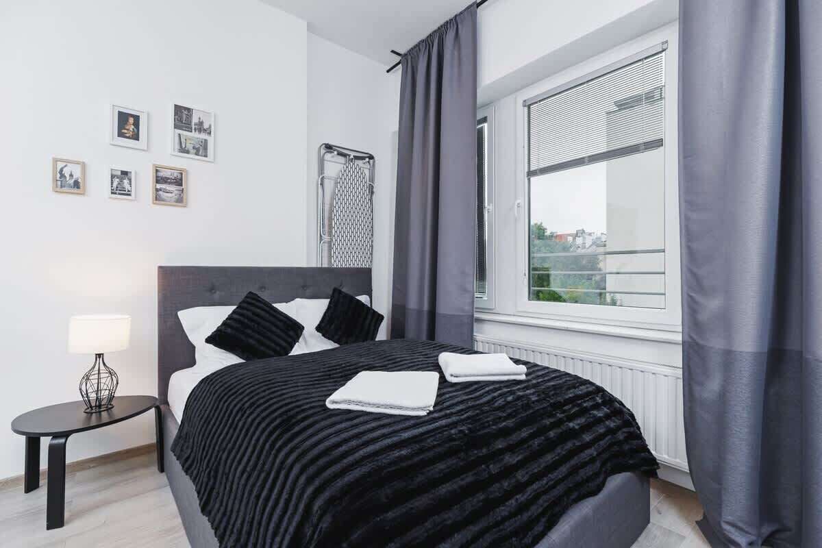 A modern bedroom with a large bed, a bedside table with a lamp, a window with curtains, and framed pictures on the wall.