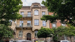 Ein mehrstöckiges Gebäude mit einem Balkon im zweiten Stock, geparkten Autos davor und Bäumen ringsherum.