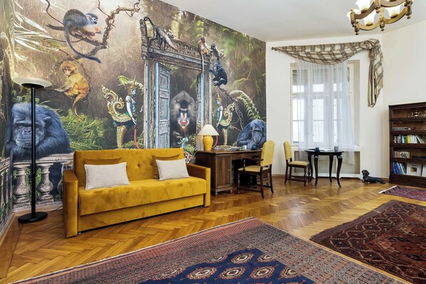 Una habitación con piso de madera estampada tipo espiga, un sofá amarillo, un escritorio con lámpara, una estantería y un gran mural con temática de selva en la pared.