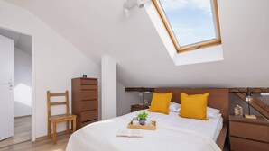 Een slaapkamer met dakraam, een bed met wit beddengoed, twee gele kussens, een nachtkastje met lamp en een houten stoel.