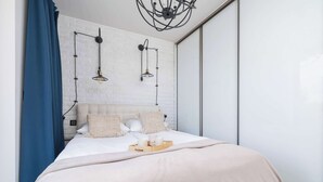 A modern bedroom with a white brick wall, a bed with a beige blanket, two bedside lamps, and a wooden tray with cups.