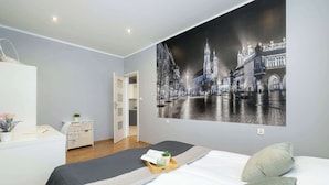 A bedroom with a large cityscape mural, a bed with a gray and white quilt, a bedside table with a plant, and a wooden tray with a book and a small plant. - Bright Apartment in Krakow | Close to Market Square, Place to Work (Kraków)