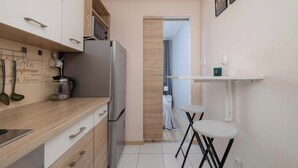 Una cocina moderna con un refrigerador de acero inoxidable, armarios de madera, una cafetera y un pequeño comedor con una mesa blanca y taburetes negros.