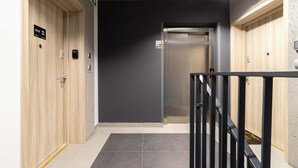 Un ascenseur moderne avec une finition métallique, une porte en bois sur le côté gauche et un mur noir portant un panneau d’indication de sécurité incendie.