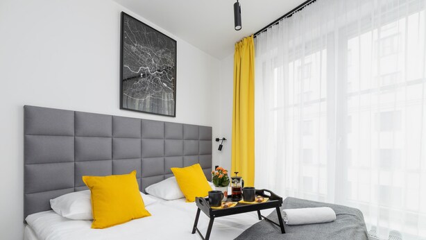 Une chambre d’hôtel moderne avec une tête de lit rembourrée de couleur grise, une grande fenêtre avec des rideaux légers et une petite table sur roulettes avec des articles pour le petit-déjeuner.