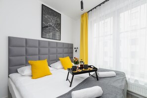 A modern hotel room with a grey upholstered headboard, a large window with sheer curtains, and a tray table with breakfast items.