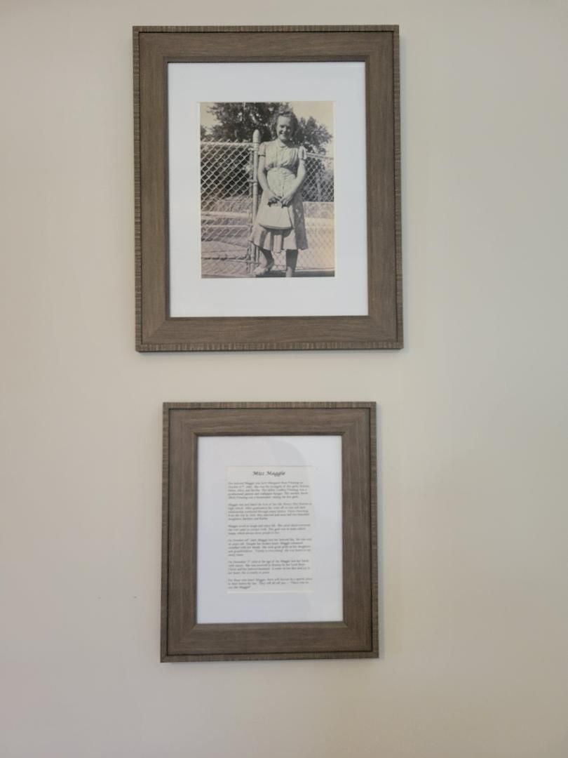 Zwei gerahmte Gegenstände an einer Wand: eines ist ein Foto von einer Person vor einem Zaun, das andere ein gerahmtes Textdokument.
