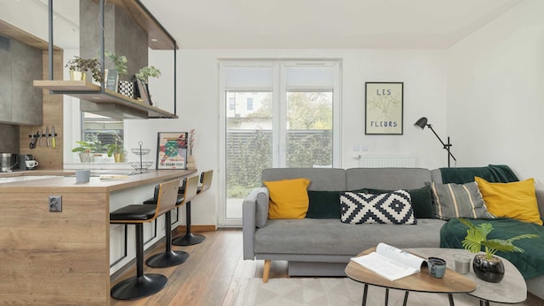 A modern living room with a grey sofa, yellow and black cushions, a coffee table, and a kitchen area with bar stools.