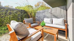 An outdoor seating area with wooden furniture and cushions, a small table with a plant, and a view of a garden and trees.