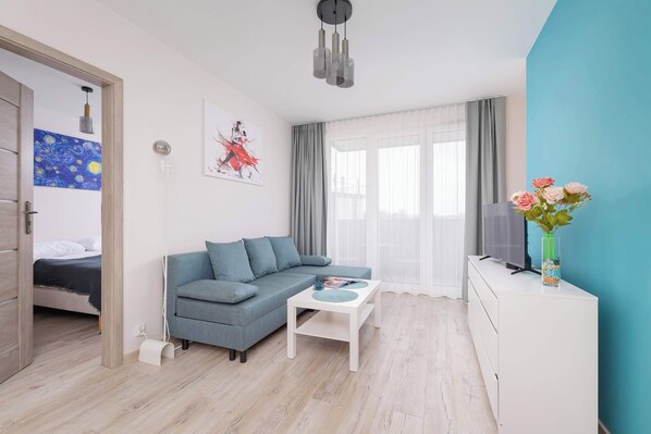 En moderne stue med en grå sofa, et lille hvidt bord og et tv på et hvidt skab. Et billede hænger på væggen, og på skabet står en vase med blomster.
