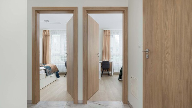 A hotel room with a bed, a desk, and a chair. There is a wooden door and a window with curtains.