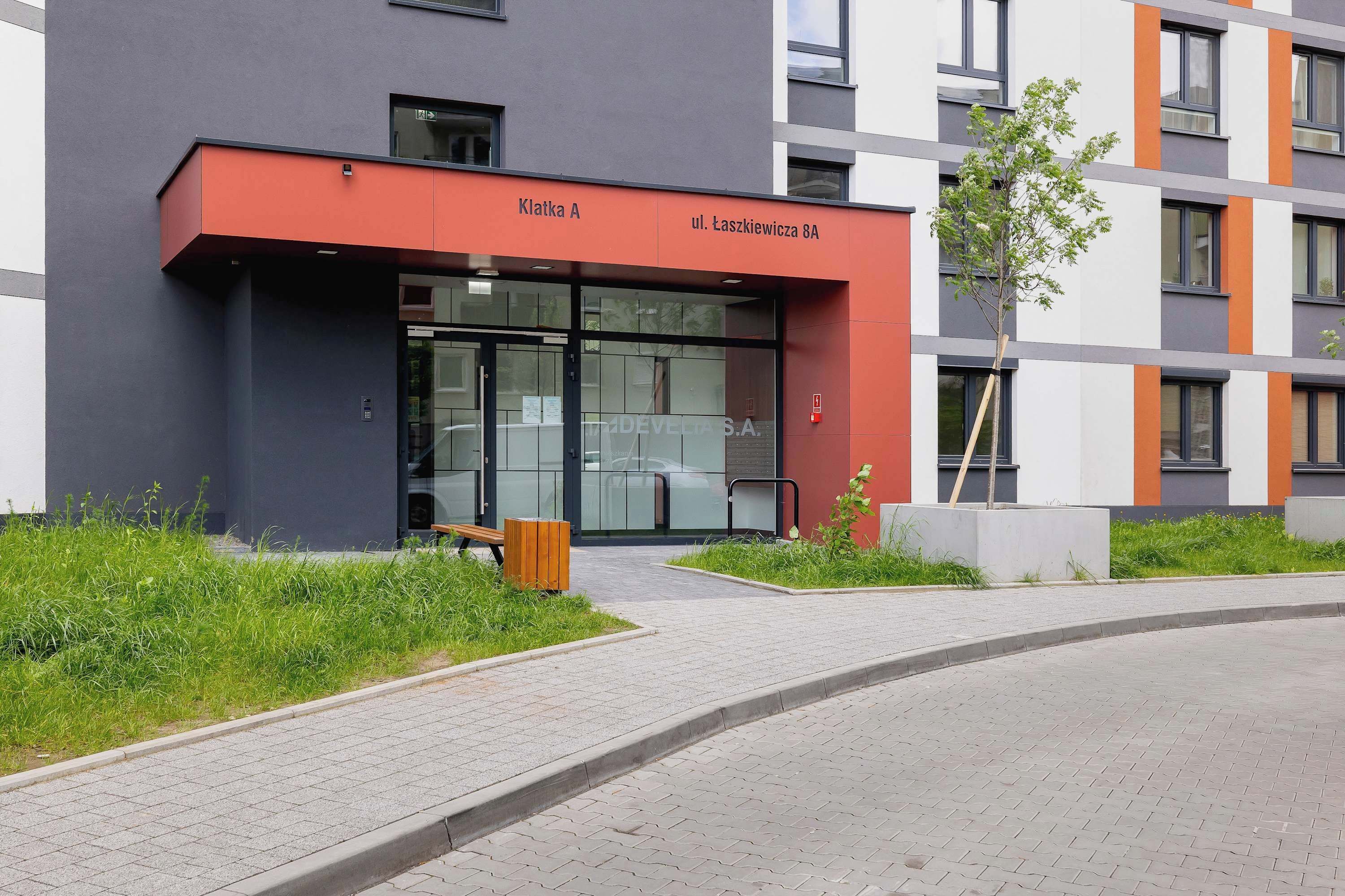 Ein modernes Gebäude mit roter und grauer Fassade, einem Glasvordereingang und einem kleinen Baum im Vordergrund.