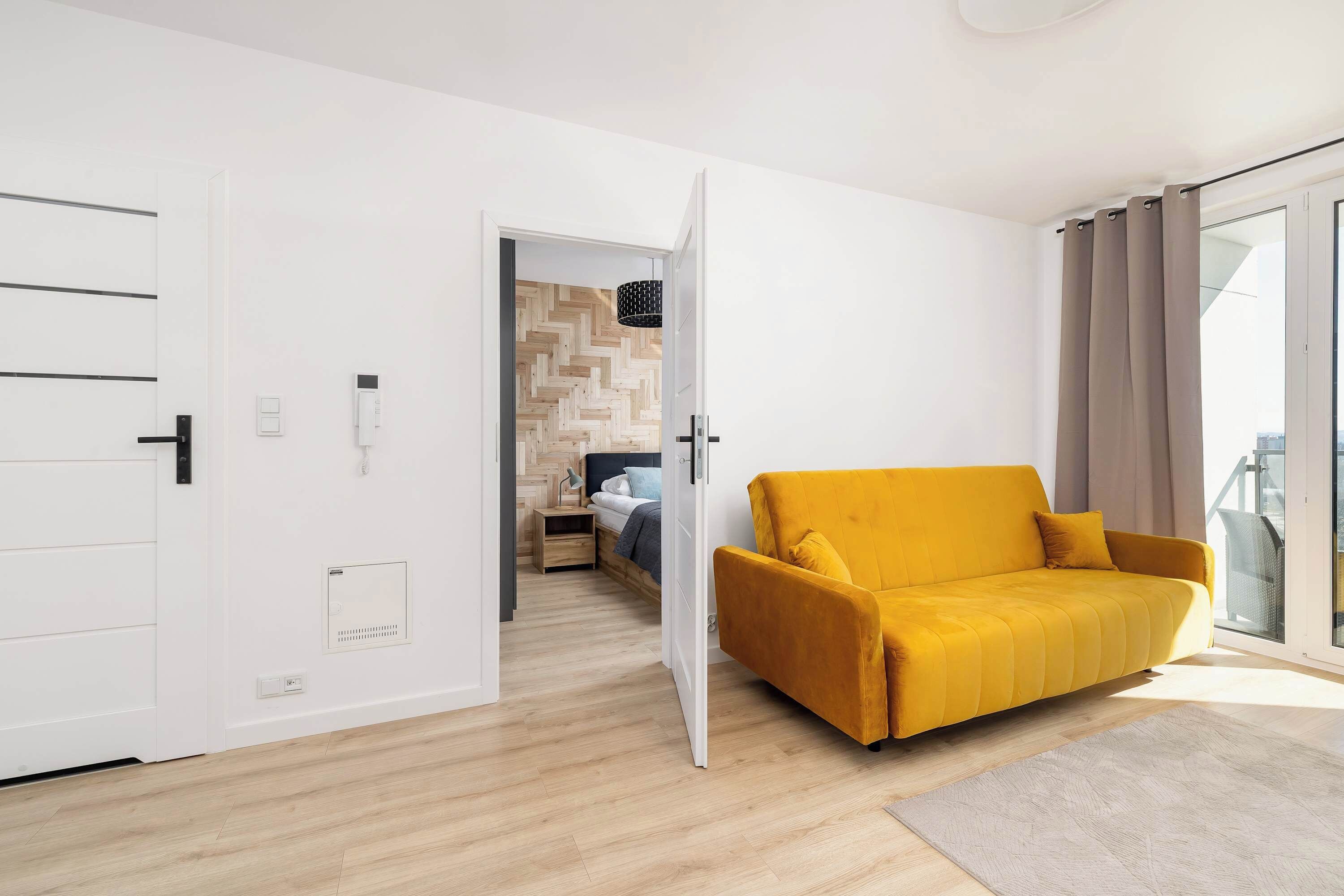 A modern living room with a yellow sofa, a wooden herringbone floor, and a glass door leading to a balcony.