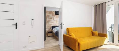 A modern living room with a yellow sofa, a wooden herringbone floor, and a glass door leading to a balcony.