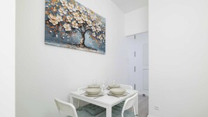 Un comedor con una mesa blanca dispuesta para cuatro personas, sillas con cojines verdes y un cuadro grande con motivos florales en la pared.