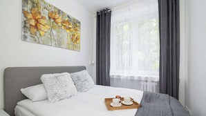 Una cama bien hecha con cabecera gris, ropa de cama blanca y almohadas mullidas. Un reposabebidas de madera con dos tazas y un pequeño recipiente se encuentra sobre la cama.