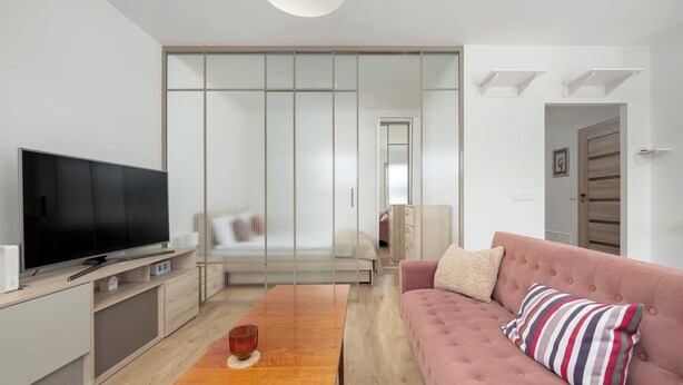Ett modernt vardagsrum med en platt-TV, ett soffbord i trä, en rosa soffa och en skjutdörr i glas som leder till ett sovrum.