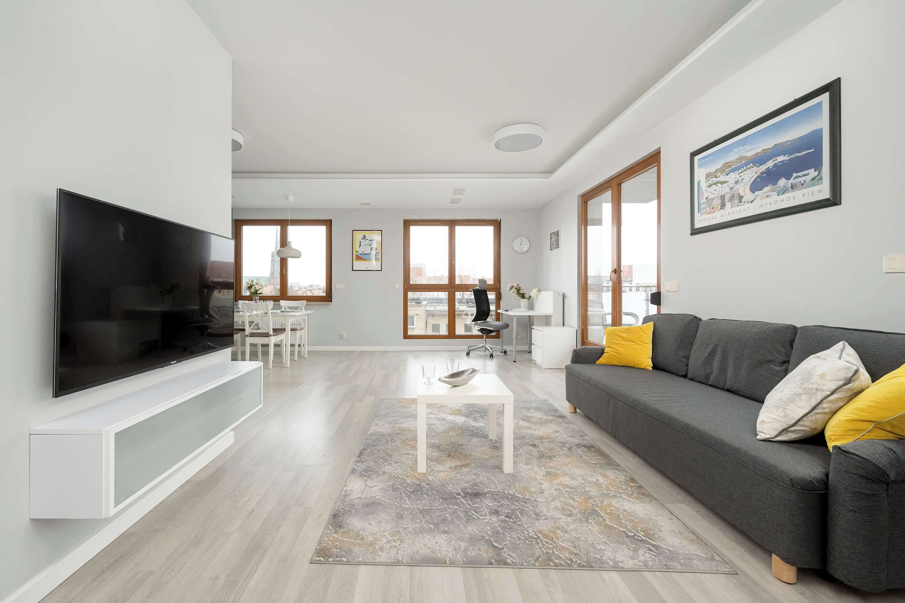 Una sala de estar moderna con un televisor de pantalla plana, sofá gris y un comedor al fondo.