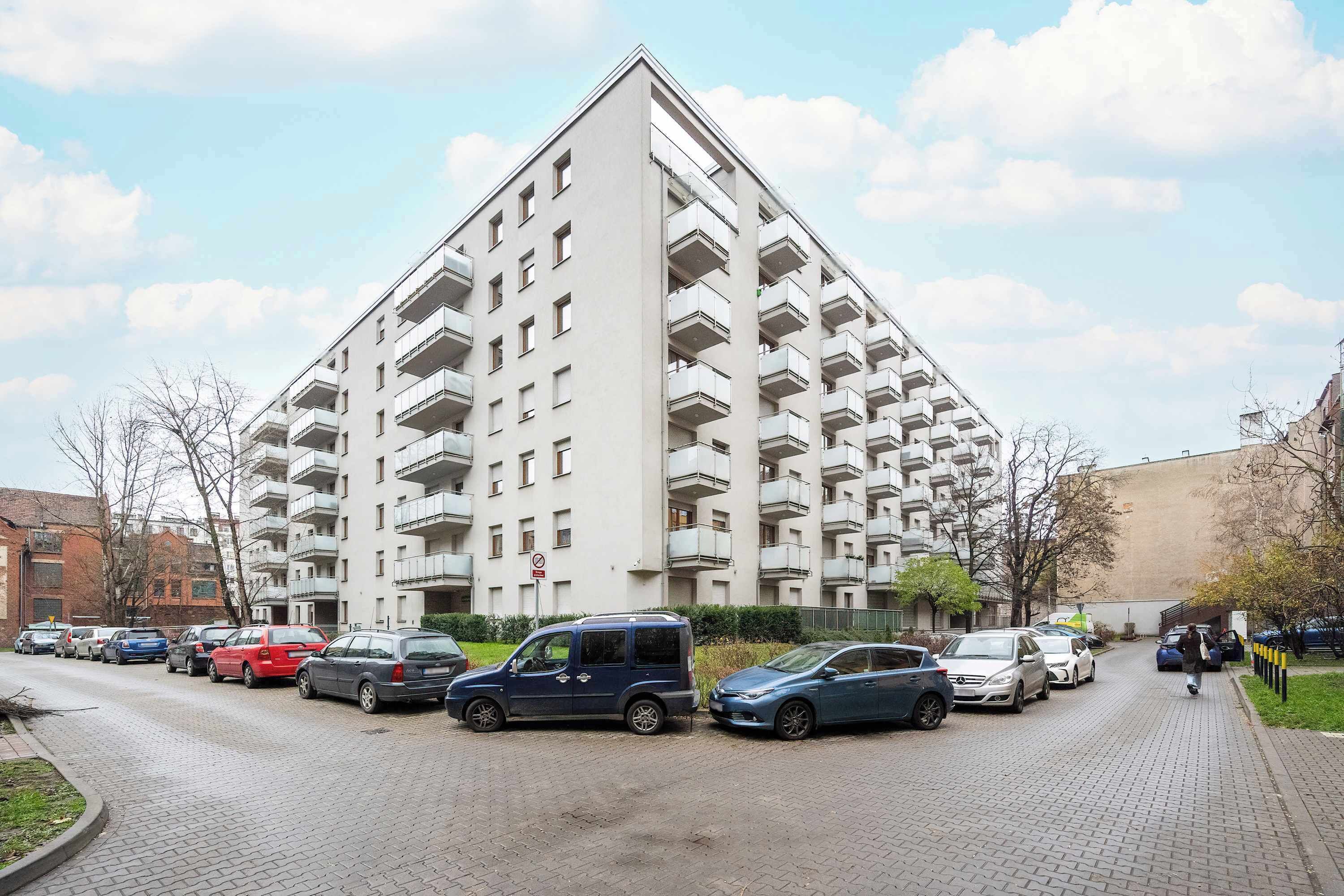 En flereetagers boligbygning med altaner, parkerede biler og en klar himmel.