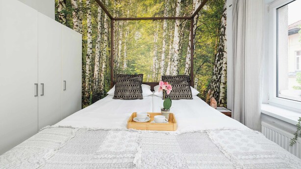 Un dormitorio con cama con dosel, ropa de cama blanca, una bandeja con tazas y una flor, y papel tapiz con motivos de bosque.