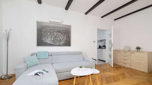 Una sala de estar moderna con un sofá gris, una mesa de centro blanca y un suelo de madera con estampado de espiga. Un cuadro de un paisaje urbano en blanco y negro adorna la pared, y un mueble con cajones se encuentra al fondo.