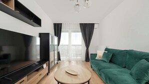A modern living room with a flat-screen TV, a green sofa, a round coffee table, and a large window with curtains.