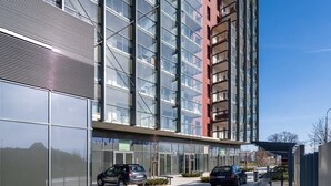 Un edificio moderno con ventanas amplias, un área pavimentada con un banco y coches estacionados en la parte delantera.