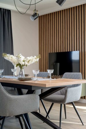A modern dining area with a wooden table, grey upholstered chairs, and a vase of white flowers. - Stylish Apartment in Wrocław Breweries | River View | Parking (Wrocław)