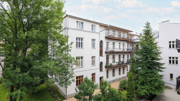 A multi-story building with balconies, surrounded by trees and a paved courtyard.