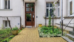 Una entrada de edificio con una puerta de madera, un pequeño jardín con plantas en macetas y una cerca de metal.