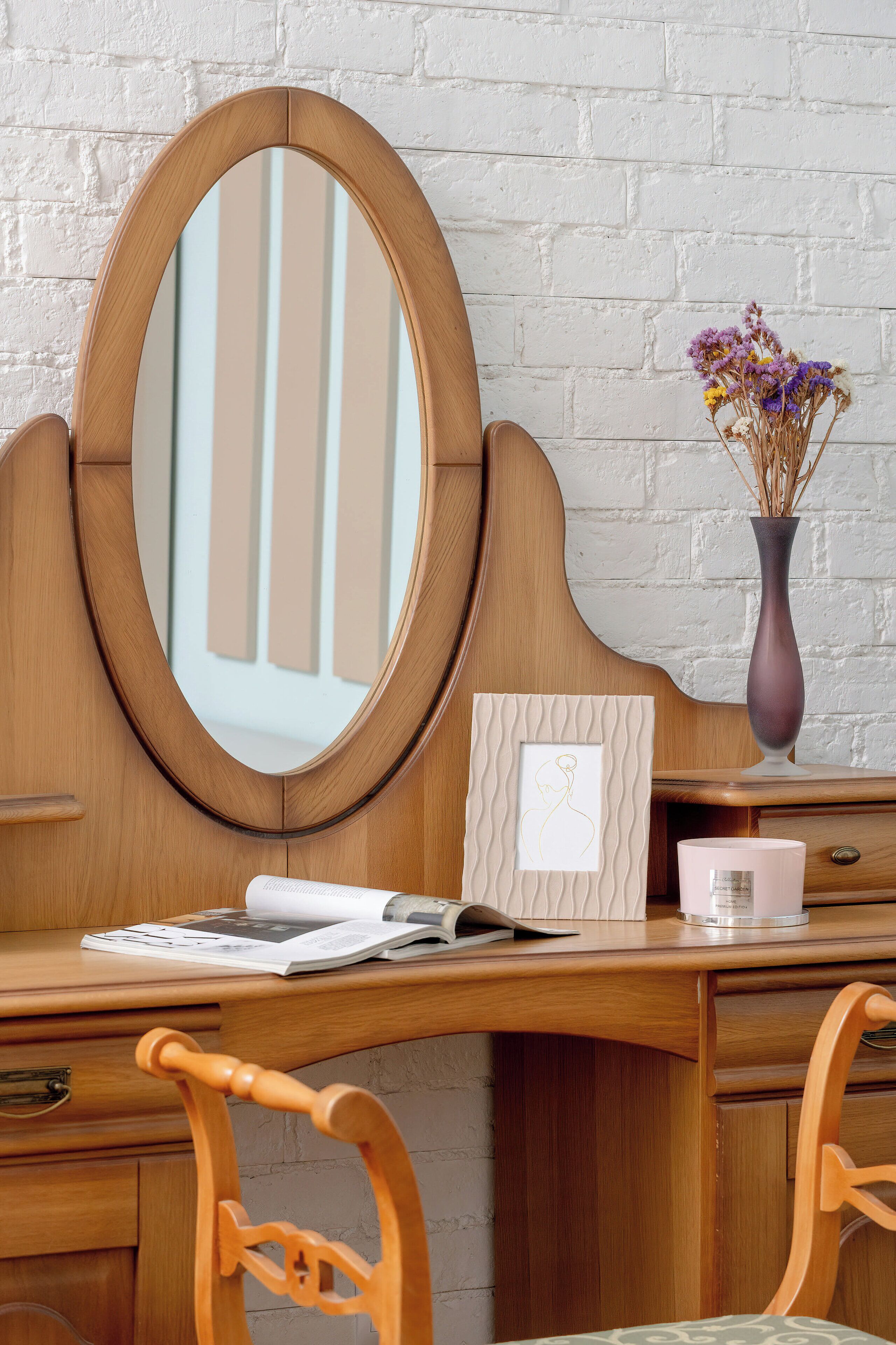 Een houten bureau met een spiegel, een vaas met gedroogde bloemen en een lijst met een foto aan de muur.