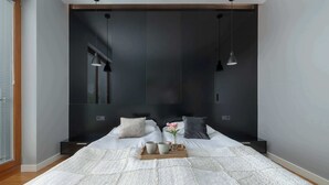 A modern bedroom with a large bed, a bedside table with a tray of cups and a vase, and a window with blinds.