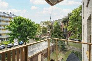 Een balkon met uitzicht op een stadsbeeld, waaronder een kerktoren, geparkeerde auto's en groen.
