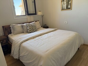 Un lit bien fait, avec une couette blanche et des oreillers à motifs, une tête de lit en bois, une lampe de chevet et un tableau encadré au mur.