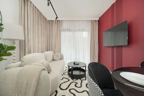 A modern hotel room with a sofa, a dining table, a TV mounted on the wall, and a large window with curtains.