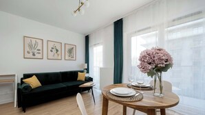 Un salon moderne avec un canapé vert foncé, une table de salle à manger ronde en bois et une chaise blanche, ainsi qu’un vase rempli de fleurs roses.