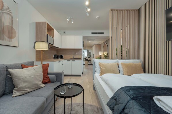 A modern hotel room with a sofa, a bed, a small table, and a kitchen area in the background.