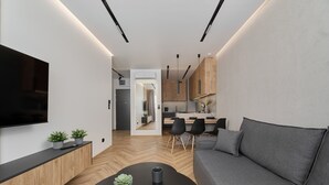 Una sala moderna con un televisor de pantalla plana, un sofá gris, una mesa de centro negra y una cocina al fondo.