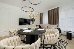 Een moderne eethoek met een ronde tafel voor vier personen, omringd door gestoffeerde stoelen. Op de tafel staat een vaas met bloemen en aan de muur is een flatscreen televisie bevestigd.