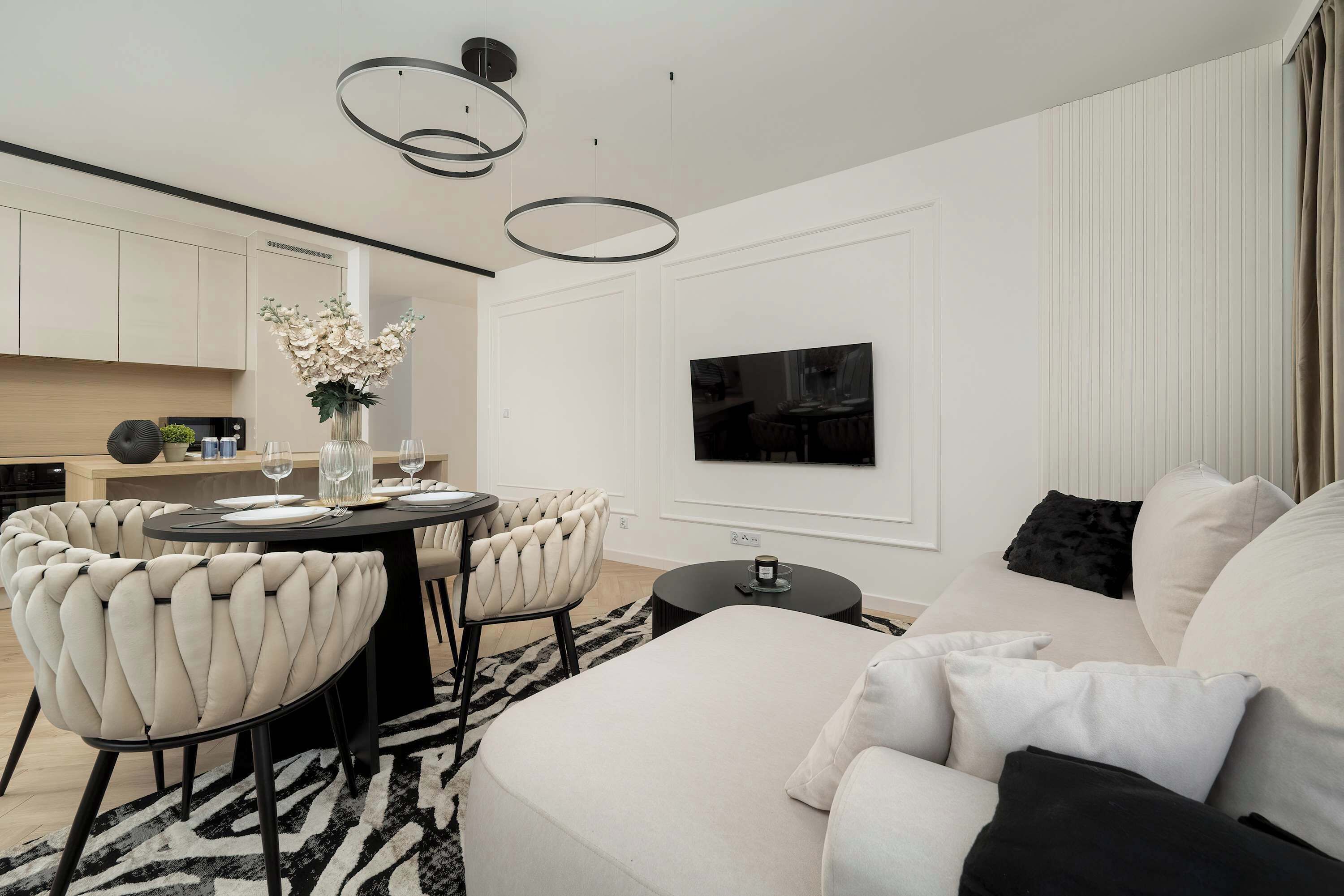 A modern living room with a sofa, a dining table with chairs, a TV mounted on the wall, and a kitchen area in the background.