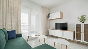 Una sala de estar moderna con un sofá verde, una mesa de centro blanca, un televisor sobre un mueble blanco y una planta en el mueble.