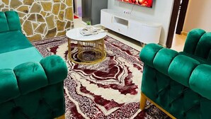 Ein Wohnzimmer mit grünen Sofas, einem gemusterten Teppich, einem weißen Couchtisch, einem Fernseher, der ein Feuer zeigt, und einer Steinwand.