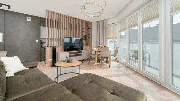 Una sala de estar moderna con sofá, televisor, comedor y ventanas amplias.