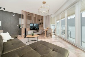 Una sala de estar moderna con sofá, televisor, comedor y ventanas amplias.