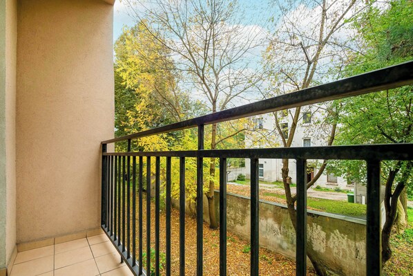 A balcony with a black metal railing, a tiled floor, and a view of trees and a building in the background.