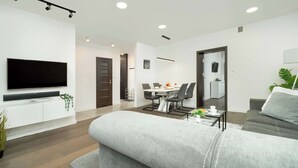 A modern living room with a flat-screen TV, grey sofa, dining area with a table and chairs, and a small side table with a cup.