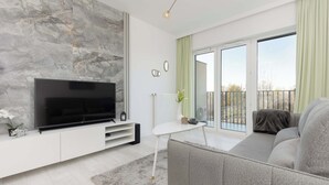 Ein modernes Wohnzimmer mit Fernseher, grauem Sofa, weißem Couchtisch und einem großen Fenster mit grünen Vorhängen.