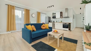 Una sala de estar moderna con un sofá azul, cojines amarillos, una mesa de centro de madera con fruta y una cocina al fondo.