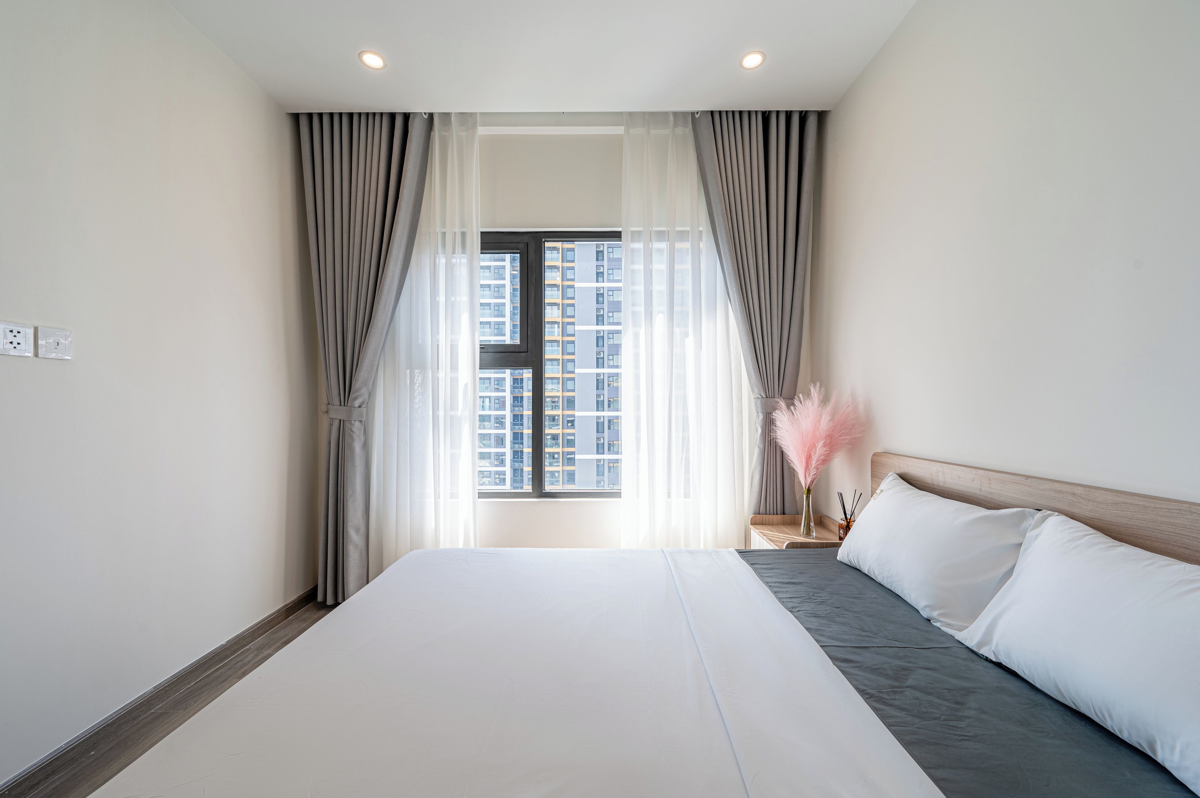 A hotel room with a large bed, a nightstand, a window with curtains, and a view of high-rise buildings.